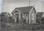 En stuga, två män och två kvinnor i Kalmar södra koloniområde, fotograferat omkring 1930. Kalmar södra koloniförening grundades 1917 och har idag 105 kolonilotter. Området ligger strax söder om länssjukhuset i Kalmar med huvudingång från Stensbergsvägen.