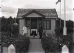 En stuga, man, kvinna och hund i Kalmar södra koloniområde, fotograferat omkring 1930. Kalmar södra koloniförening grundades 1917 och har idag 105 kolonilotter. Området ligger strax söder om länssjukhuset i Kalmar med huvudingång från Stensbergsvägen.