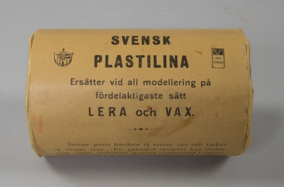 Plastelinamaterial, 6 st. 4 st cylindriska i obrutna förpackningar med brunt omslagspapper och lång reklamtext. 2 st utan omslagspapper och använda till häften. Ovanpå en av dem klumpar av plastelina.