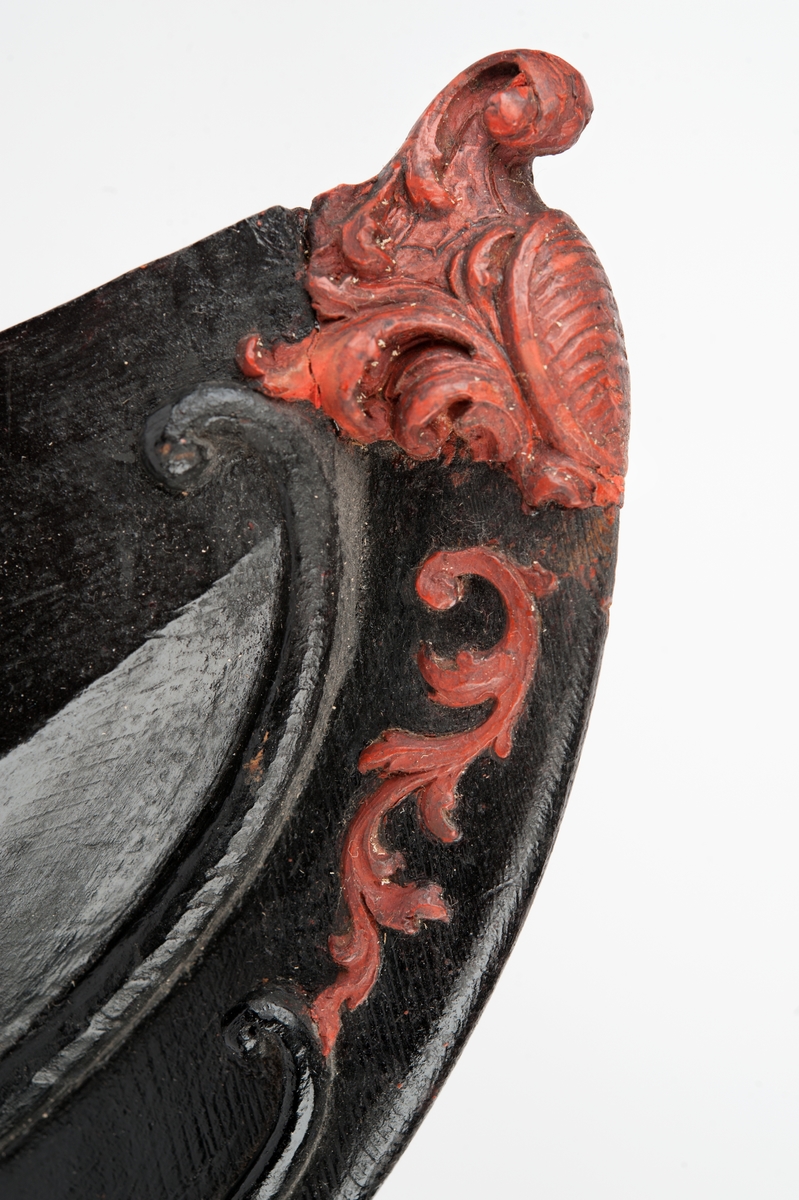 Okänt fartyg. Vapensköld med räfflad botten, krönt av volut och omgiven av blad och bladslingor. Monterad till stävkonstruktion av svartmålat trä. Bottenplattan saknas. Röd vax.