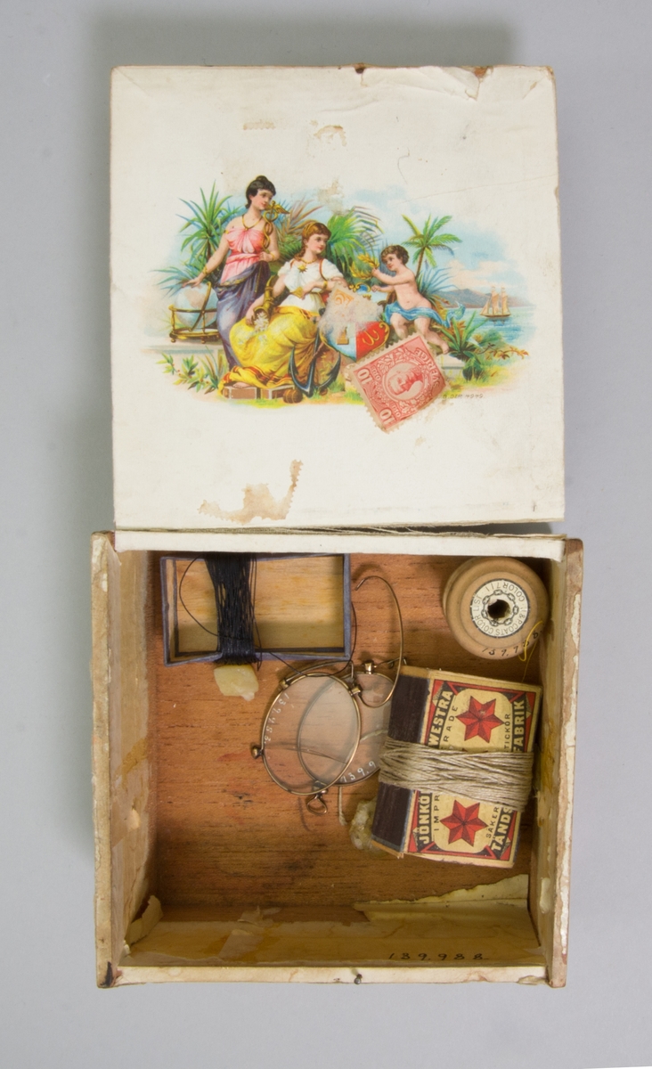 Sydonsask, fd cigarrlåda, av trä kvadratisk med fällock. Innehåller bland annat en rulle med gul tråd, en del av en tändsticksask omlindad med svart tråd, en del av en tändsticksask omlindad med beige tråd och två synålar samt trasig lornjett med guldfärgade bågar. På lockets insida påklistrat frimärke 10 öre med Gustav V:s bild, rött.