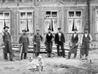 Män i förkläden med sågar och andra verktyg i händerna, stående framför ett husbygge, troligtvis ett hus för arbetare i Norrsundet. Mannen i väst och kavaj kanske är förman. Två barn sitter på marken.