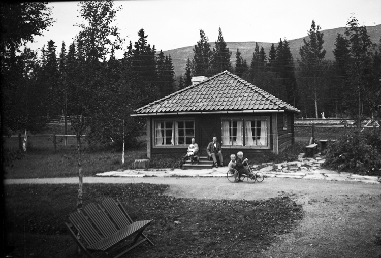 Framför ett bostadshus (?) sitter några vuxna och två pojkar sitter i en leksabil.                                                                                                                                                                                              
I förgrunden syns en trädgårdsmöbel.