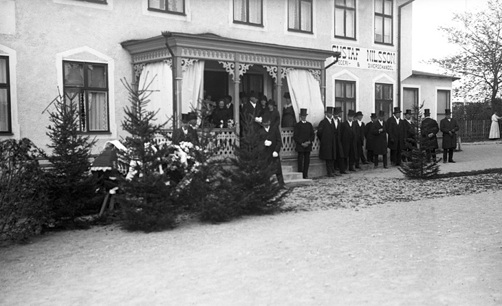 På bilden syns ett antal personer framför en affär/bostadshus i samband med en begravning.
Trol begravning (maj 1919) av fru Ester Sigrid Kristina Nilsson (1884-1919), Braås. Gift med handlande Gustaf Nilsson (1870-    ), Braås.
Källa: Sjösås Dödbok 1919.