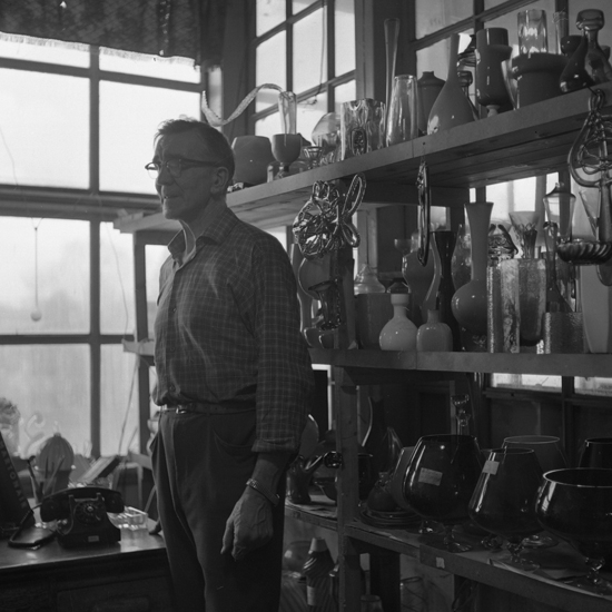 Foto från arbetet i en glashytta. Glasarbetare framför hylla full med glasföremål, vaser m.m.