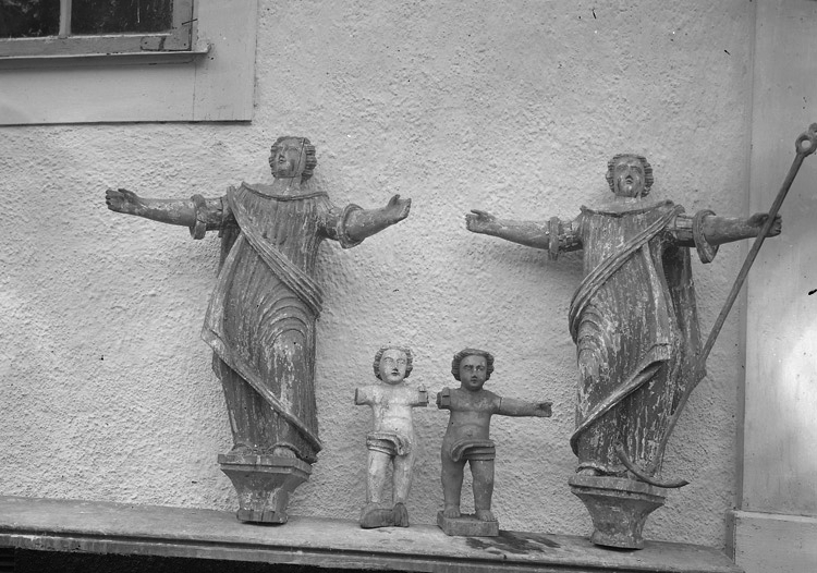 Foto på en bänk med kyrkliga träfigurer (antagl. från altaruppsats) utanför kyrkan.