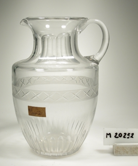 Kanna, 100 cl.
Tillverkad på Sandvik-Orrefors glasbruk 1910-1942.
Form inspirerad av antiken.
Matt bård med kryss, slipade oliver längst ner och på halsen.
Något dragen pip, påklipp till grep.
Driven och kulad i botten.
Klarglas.
Lapp "N.K 100 cl."
Gåva av Orrefors glasbruk år 1968.
Funktion: Kanna för vatten el. likn.