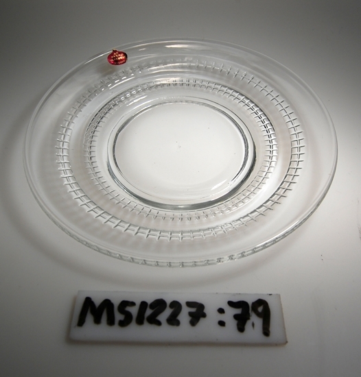 Assiett av sodaglas.
Rund assiett. Brättet har två stycken horisontellt löpande linjer med korta vertikala linjer.
Klarglas.

Inskrivet i huvudbok 2011.
Funktion: Assiett