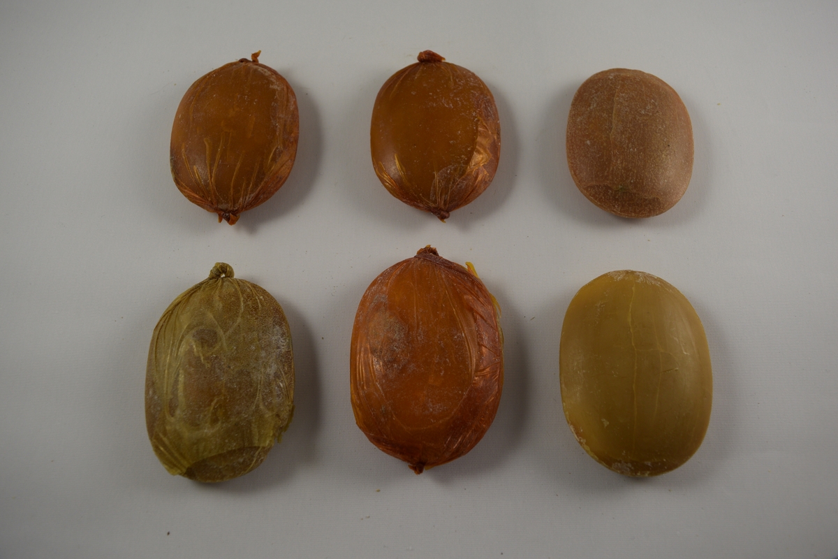 Oval såpe, seks stykker som er tilnærmet like. Produsert ved Saanums Sepefabrikk i Mandal. Tre stykker pakket i oransje plastikk (a-c), en i brun plastikk (d). To uten plastemballasje (e-f), men i ulik farge. Alle dekorert med Mandals kommunevåpen, med tre laks.