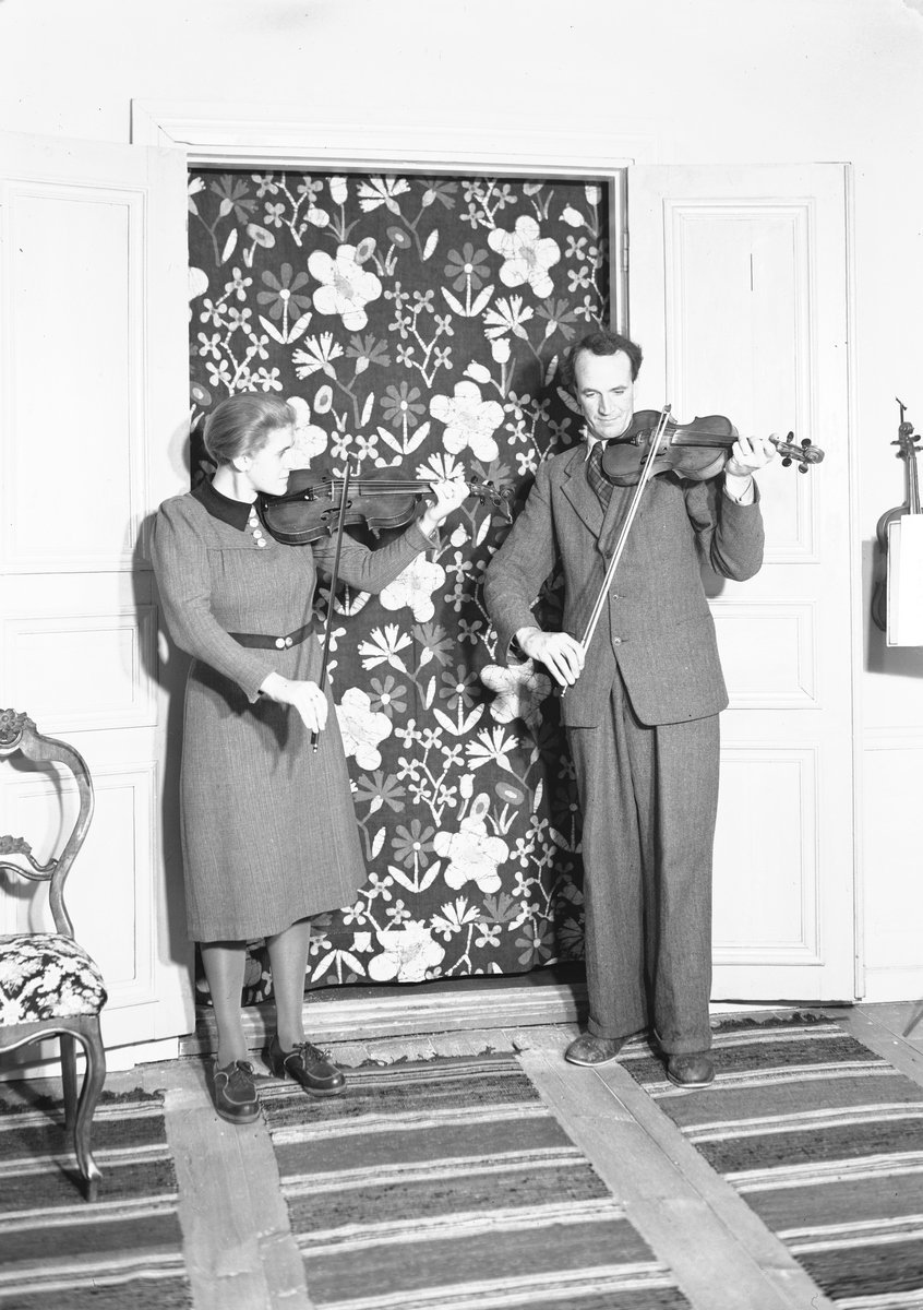 Manne och Brita Östlund hälsar gästerna välkomna med Jons-låten. Bakom dem ett batikdraperi utfört av Brita med röda, vita och blå blommor på violett botten. För "Svenska Hem i ord och bild"
