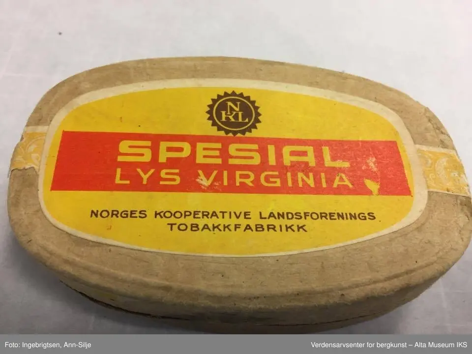 Ovalformet eske med innhold av tobakk og papir. På lokket er det et klistremerke med logoen til Norges Kooperative Landsforening (NKL) og navnet på produktet. På undersiden av esken er det ei remse som er klistret på med informasjon.