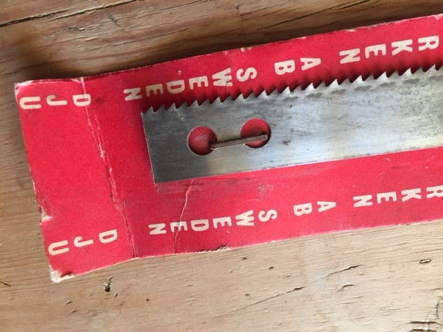 Ubrukt sagblad montert på originalt forpakning bestående av en rektangulær papp-plate. I enden av papp-platen er festet en hyssingstump for opphenging på vegg. Trolig er bladet beregnet til en grindsag.