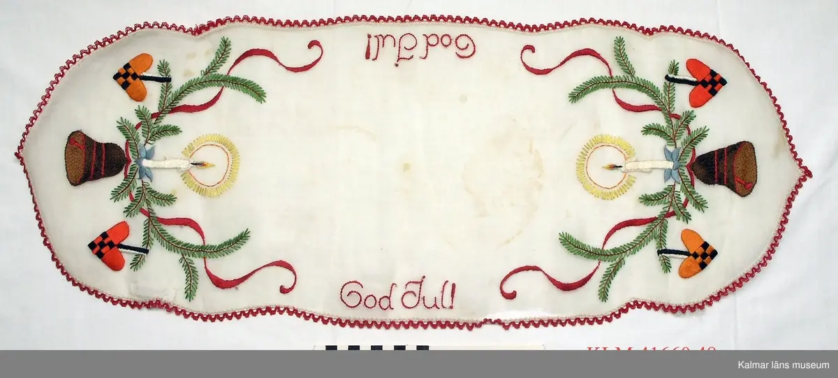 KLM 41660:48 Julduk, textil. Avlång, vitt tyg med mönster i fritt broderi i rött, grönt m fl färger, grankvistar med hängande hjärtan och klocka, ljus samt texten "God Jul!", smal röd spets runt om.