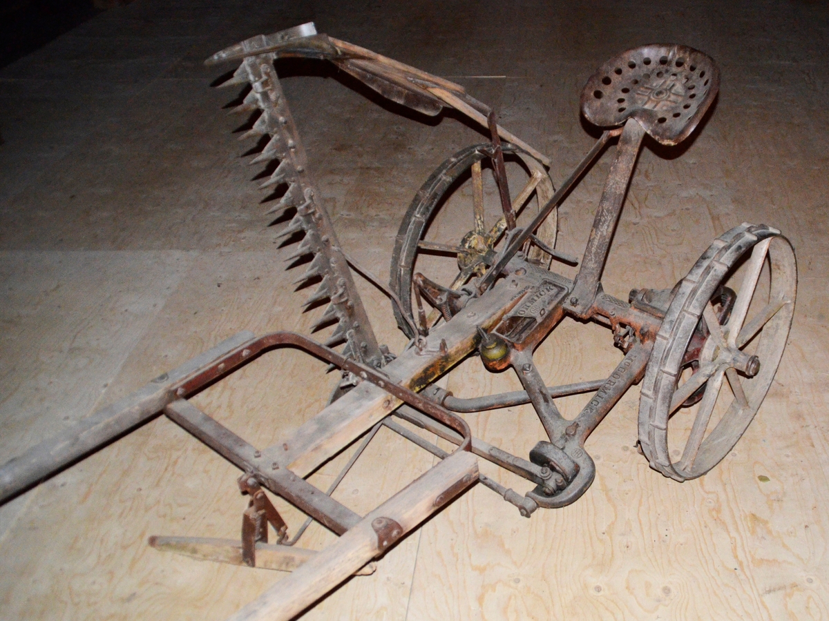 Slåmaskin for hest med drag. 6-eika metallhjul. Sete. Jarn, utan draget, som er i tre.