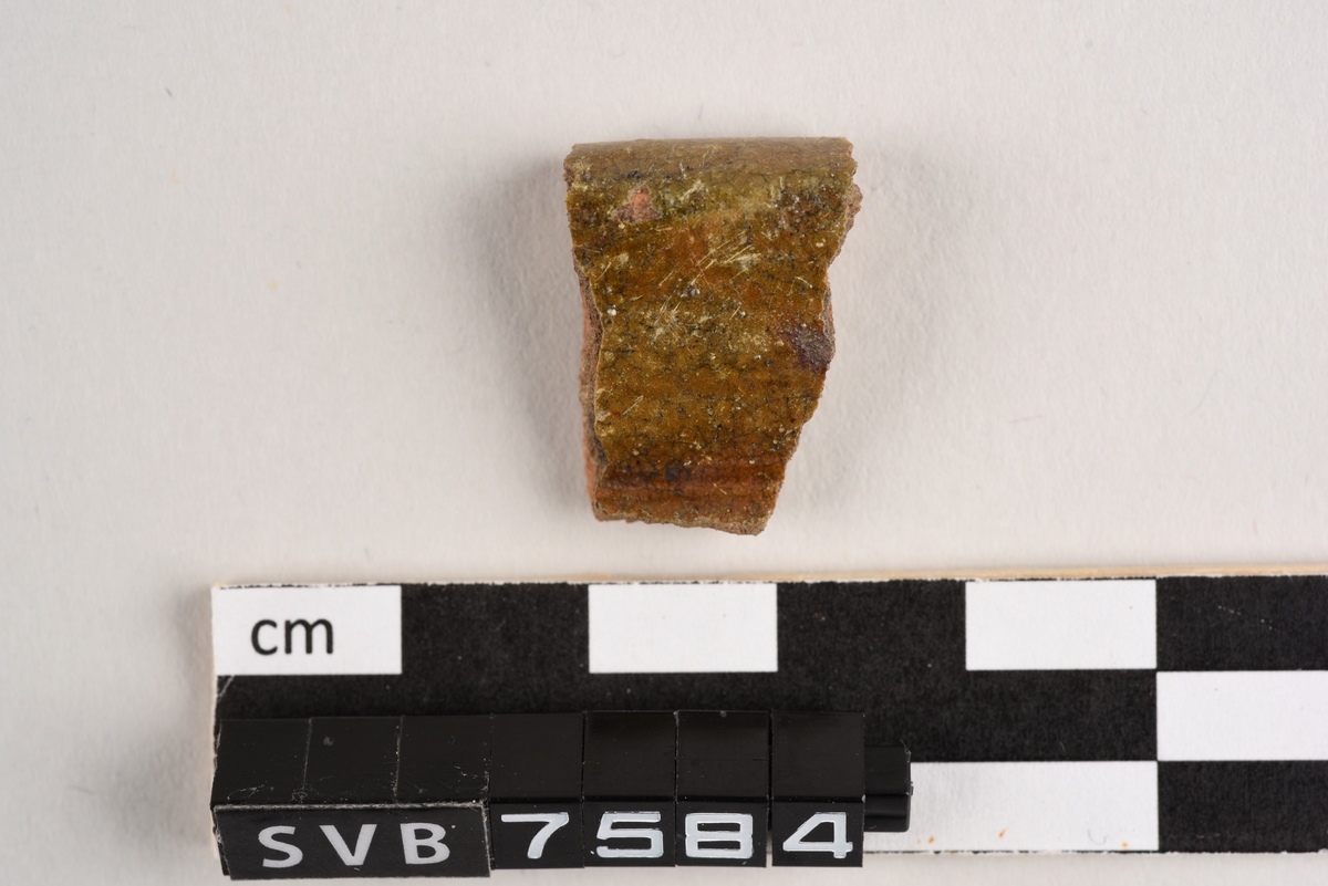 Ett rødlig keramikkfragment. Fragmentet er et randskår og har rester av brun glasur på begge sider.