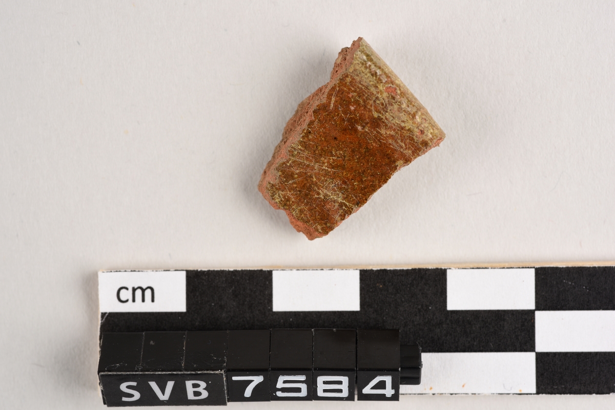 Ett rødlig keramikkfragment. Fragmentet er et randskår og har rester av brun glasur på begge sider.