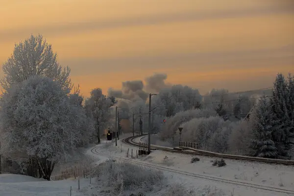 Damplokomotiv i vinterlandskap i skumringen. Himmelen i bakgrunnen er sløret og gyllen.