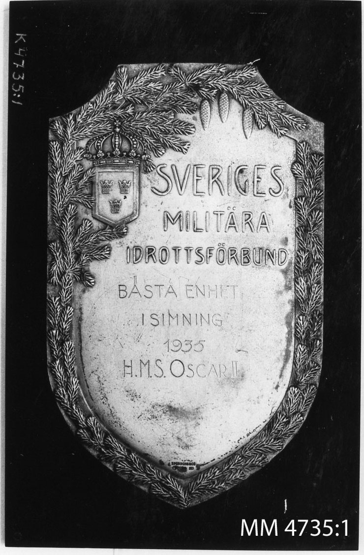 Plakett av silver fastsatt på en svart platta av ebonit. Den utgör pris i simning.
På skölden text i relief: Sveriges Militära Idrottsförbund. Ingravering: Bästa enhet i simning 1935 H.M.S. Oscar II.