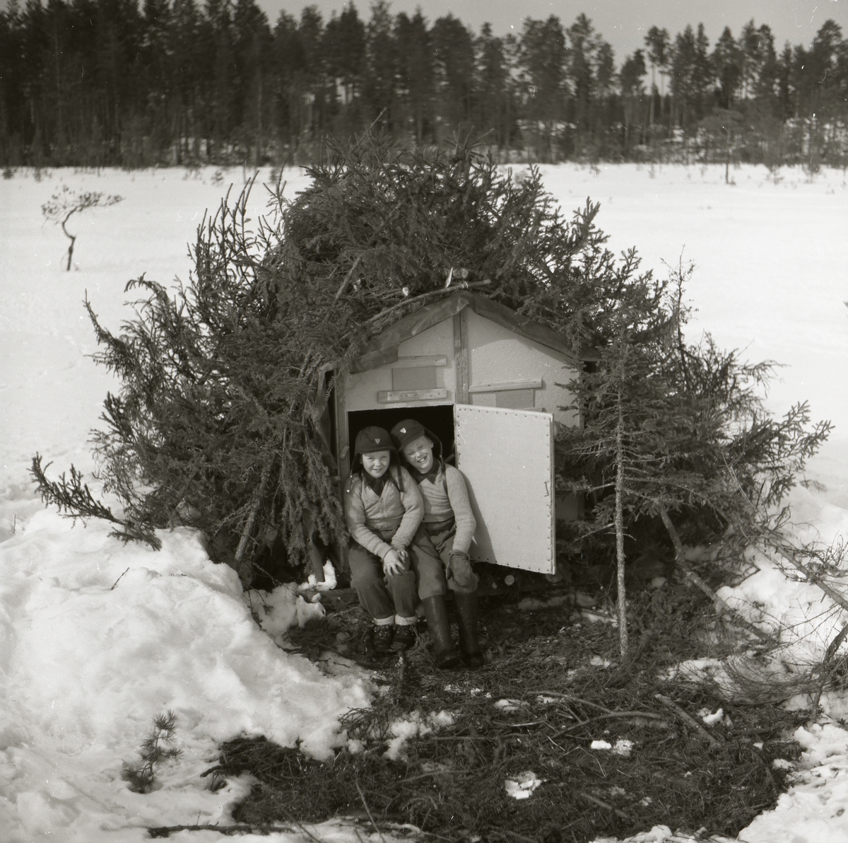 I dörröppningen till en koja sitter två barn och ler mot fotografen. Pojkarna bär matchande kläder bestående av ljusa tröjor, gråa byxor och svarta mössor. Kojan täcks av granris över taket och framför dörren ligger en granrismatta. Runt kojan ligger snön decimeterdjup med enstaka fotspår som avslöjar människornas närvaro. Skogen i bakgrunden ramar in det öppna fältet där kojan är placerad.