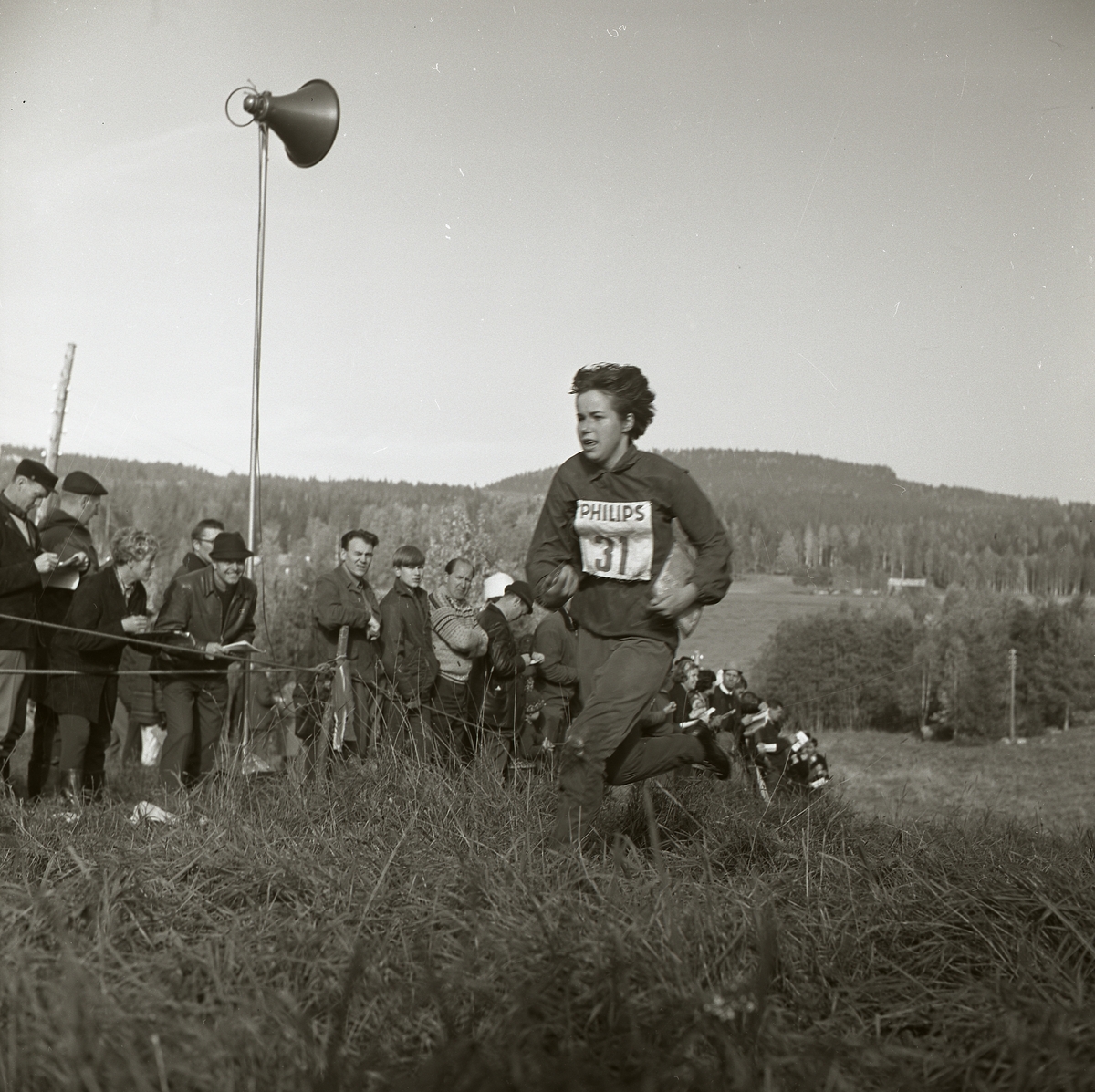 En kvinna springer iförd nummerlapp upp för en backe. Hon bär på en karta och är deltagare i svenska mästerskapen i orientering som hålls i Segersta 1965. Till vänster har en publik samlats bakom en avspärrning. På en stolpe sitter en högtalare som är placerad mitt i folkhopen.
