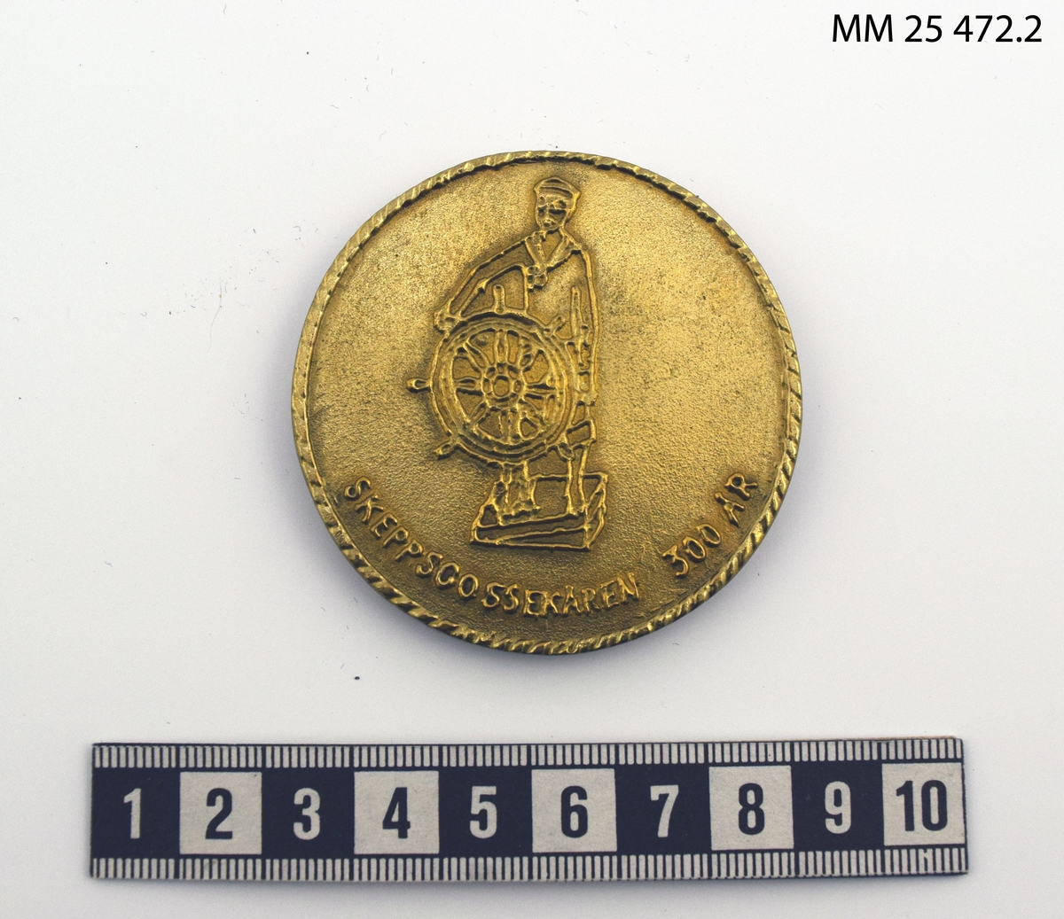 Rund plakett av gulmetall med präglat motiv. På ena sidan motiv av skeppsgosse vid styrratt samt texten: Skeppsgossekåren 300 år". På andra sidan motiv av korvetten Carlskrona samt texten: "Karlskrona 1685-1985" "Korvetten Karlskrona 1841". Plaketten förvaras i blå plastask med sammetsklädd insida.