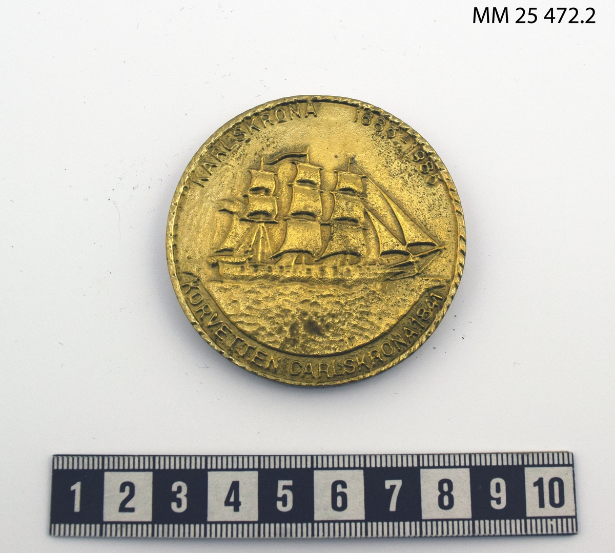 Rund plakett av gulmetall med präglat motiv. På ena sidan motiv av skeppsgosse vid styrratt samt texten: Skeppsgossekåren 300 år". På andra sidan motiv av korvetten Carlskrona samt texten: "Karlskrona 1685-1985" "Korvetten Karlskrona 1841". Plaketten förvaras i blå plastask med sammetsklädd insida.