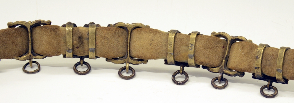 Fra protokollen: Tvislir med 1 kniv og belte av semska skinn med smøygjestolar av massing med ein nedhangande ring or kvar stol eller kvar sylgje. Beltekroken er bladforma, uthoggin (gravert) i strikmynster, fraa denne gjeng 7 taattar traadlekkje, av massing og forma som kyrmaal-taattar. Kniven med treskaft (barlind) og svære, støypte i massing - holkar (uthogne) støypte i rank eller strik. I enden paa bakholken kvardar rundt og krosslykkje i tverenden. Knivsbladet langt, med sneideslipa odd-fase i bakken. Slirine med fergd strik-ornamentik. Paa den eine innskrifti K.A.S.H, paa den andre: ANNO 1731.
Fra Telemarki eller Sæterdalen.