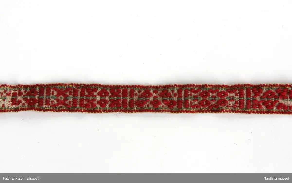 Huvudliggaren:
" 'Kass fr. Mora sn, OfvanSiljans fgd Dalarna.
Med band och sticka.
Stickan märkt: 'LHS. FÖD. 15.Mars. 1822 KHD.FÖD
INVNB 1829. AHD.9. ÅNVARI.1826. DÖD 21/6 1830
AHS:FÖD:6/10 1815 KHD.FÖD.14 ARL1820: DÖD19/41821

a. Kass.
b. Kassticka"

a: Dopkass av flaknäver med handtag. Kassen - korgen - har en långsmal form. Flera lager av näver har vikts och sytts ihop med klyvsöm. På kassens långsida finns en "krage" 6-7 cm bred. Kanten har en vidja lagd på ut och insidan av sidoväggen och gavlarna. Vidjan är fäst med en tät lindning av rottågor, i kanten följer en smal vadmalsremsa. Sidohandtagen består av en bågformad käpp, de är fastsurrade i  ändarna och i kassens övre kant. Under klyvsömmen ligger röda och blå vadmalsremsor som förstärkning och dekoration. Kassen är dekorerad med  "vadmalsnoppor"och små olikfärgade vadmalsbitar sydda på varandra. Tygbitarna är fastsydda med en lintråd som löper längs kassens insida. .Årtalet 1793 är inristat på ena gaveln.
Anm;  Trasig i ett bottenhörn. Delar av vadmalsremsor och dekoration är borta.

c: Mönstervävt band, 'krusband',  med tofsar, 'tuppor',  i ändarna. Varp av viitt bomullsgarn och rött ullgarn  med grön och gul mitt- och kantbård, inslag av bomullsgarn. Bandet har varierande mönsterformer avdelade med stolpar. I ena änden har varpen delats och avslutats med två styck fyra  cm långa tofsar, av tygremsor i bl a siden, den andra änden avslutas med en tofs.
Anm: Slitet, delar av mönstret  bortnött.

Dopkass, barnkass eller kristningskass som den också kallas, är en korg eller vagga som förekommit mest i övre Dalarna, men även i Väster- och Österdalarna och i södra Härjedalen. Den användes till dopet och andra kyrkobesök och som sovplats i hemmet. Den kunde också bäras på ryggen, då hade man ett långt band som lindades runt handtagen och sedan runt axlarna på kvinnan. Till kassen kunde höra en dopsticka, i den skar man in barnets initialer, födelsedag och år. Vid högtidliga tillfällen lindades ett mönstervävt band, såsom c,  runt handtagen på dopkassen.

En lång läderrem med smidda krokar, "dingelkrok", 12 581, användes vid upphängning av vagga, barnkorg eller dopkass. Krokarna fästes kring handtagen och  läderremmen fästes i takkrok eller dyl.
/Inga-Lill Eliasson 2006-04-28