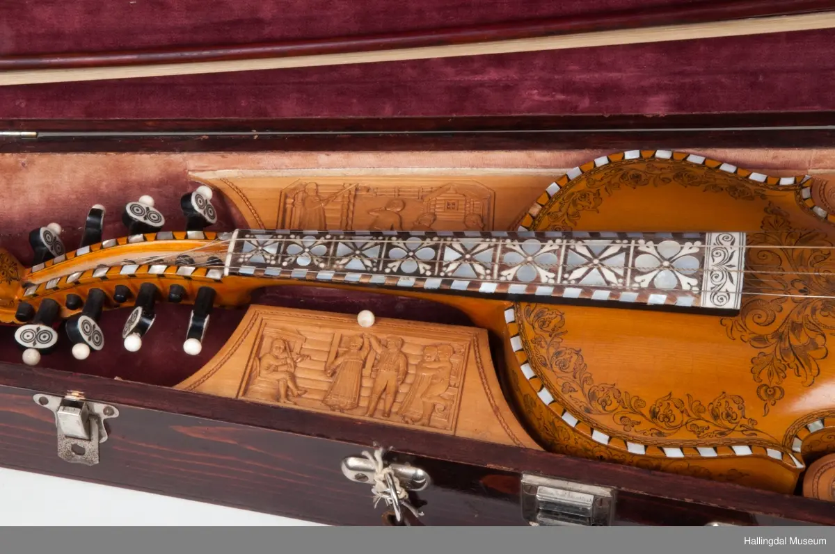 Lokket er utskjært med drake og båndslynginger.  Utskjæringer inne i felekassen.  5 rom med lokk i kassen.  3 lokk har utskjæringer med motiver fra hallingdans.  
Skaftet er besatt med perlemor, rosetegning i sort, dyrehode øverst på skaftet.  4 strenger over og 4 strenger under strengbrettet.  2 felebuer rødbrun mahogni.  Fela se HFN 03210.