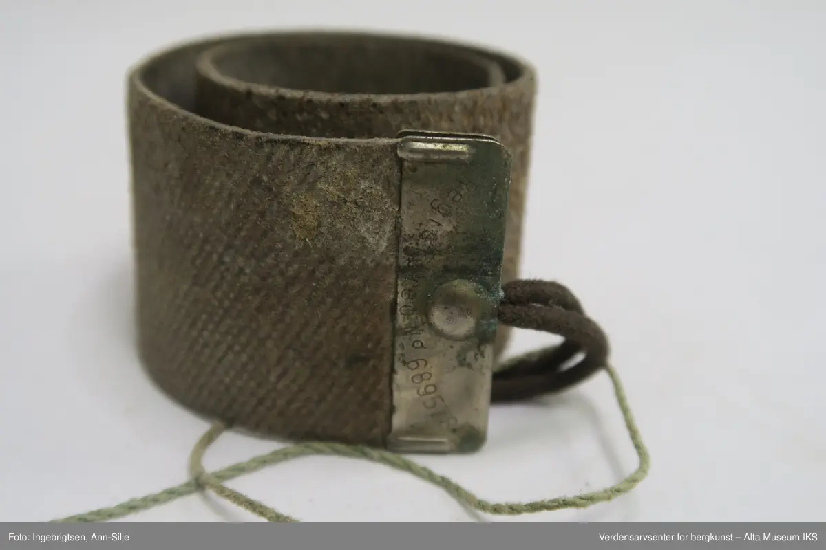 En rektangulær lærremse med et metallstykke klemt i den rette enden. Remsen ender i en spiss i den andre enden. En løkke av tau er festet i metallet. På oversiden av læret er det rutemønster i læret, mens undersiden er glatt.