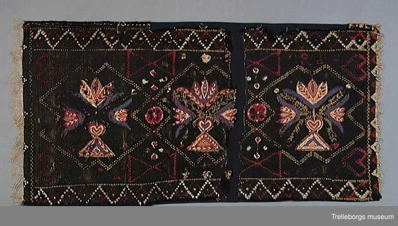 Material: varp 2 tr lingarn, inslag 1 tr ullgarn, ripsbindning. Broderi och flossaknutar 1 tr ullgarn. 
Bården runtom är vävd i tränsaflossa, diagonal inrutning. Dessutom är vävt konturer till tre rutor, dessa delar in agedynans mittfält i tre avdelningar. I tränsaflossan är även invävt konturerna till en hjärtfigur i vardera hörnet, samt två timglas utmed långsidorna. I var och en av de tre rutorna är broderat en blomstervas med tre tulpaner. Färgerna är här blått, cyklamen, gult, vitt och olivgrönt.

Tidigare konserverad då bitarna har sytts ihop på ny svart botten. Eventuellt kan det vara så att dynan klippts sönder för att brukas i två delar eller vid arvsskifte.