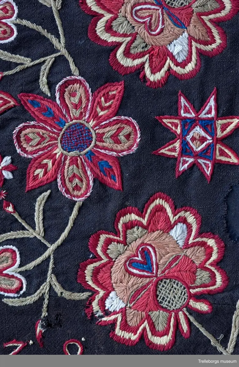 Botten är svartbrun tuskaft i ullgarn. 
Ringkrans med 4 blommor.
Hela mönstret är spegelvänt utmed en längsgående axel (frånsett 2 tulpanfigurer och märkningen ALD HMDG utmed den övre långsidan och ANNO 1838 utmed den nedre långsidan. 
Broderiet består av gängse fantasiblommor och tulpaner, omgivna av bladslingor.
Färgerna är vitt, blekgult, blekgrönt, blått, bronsgult mm