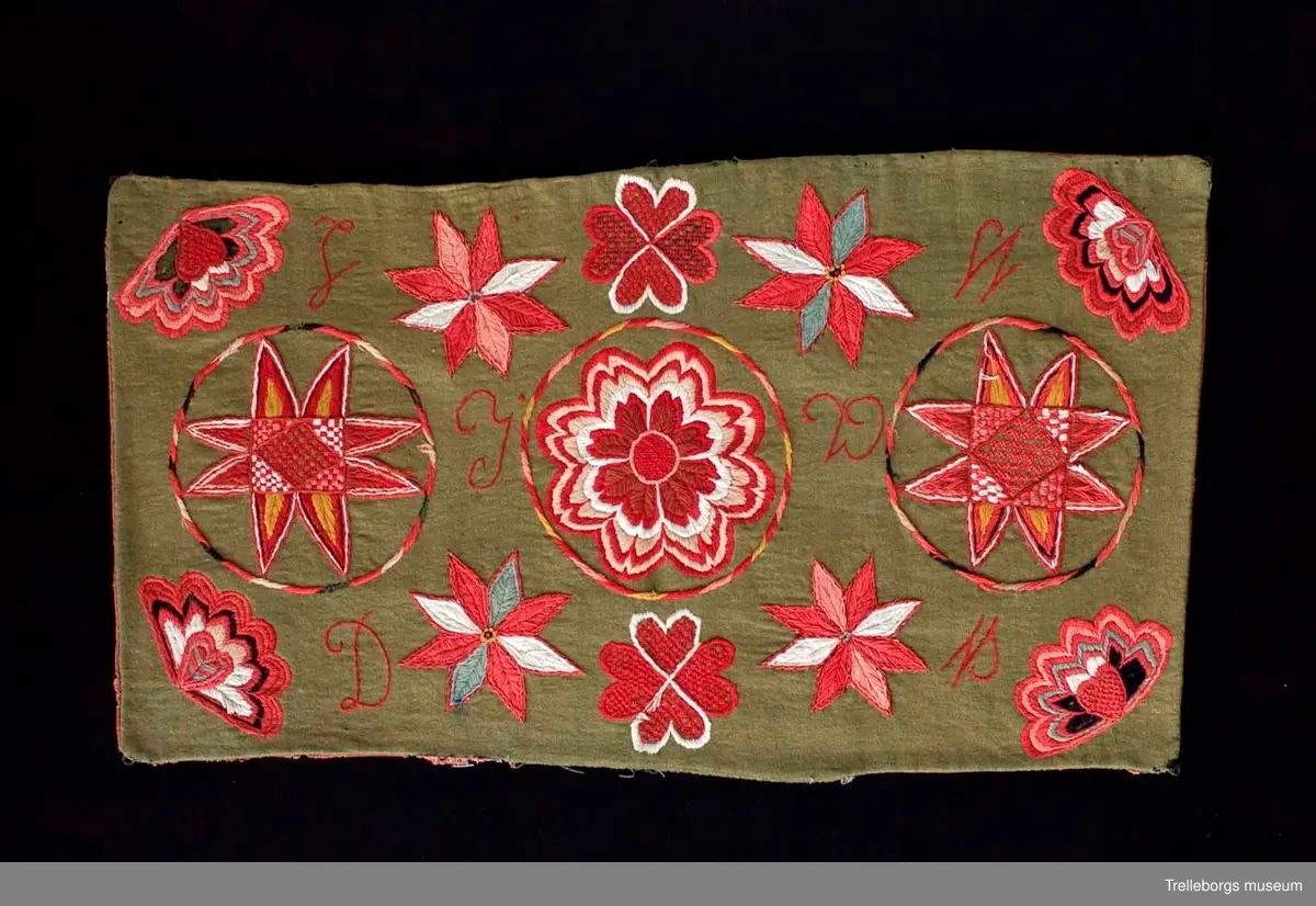 Bottentyg är grön vadmal. Tre stora ringar i kontur utgör huvudmotiv. I ringen i mitten är en stor blomma med 8 kronblad utfyllnad. I de båda ringarna vid sidan är broderat en spetsuddig stjärna med 8 vingpar. Dess centrum är en snedställd kvadrat, sydd i gallerliknande konstsöm. Utmed agedynans långsidor är broderat olika blomsterstjärnor, pionliknande blommor och en blomliknande rundning av 4 st hjärtfigurer. Färger: vitt, gult, svart, blågrått samt många olika nyanser i rött. Baksidan tegelröd yllevävnad. 
Enligt katalogen märkt I.W.D.S.  SL. Hjärtrosetter och hjärtan. 
Enligt Ernst Fischer, Årsbok från Malmö museum 1971, sid 180: En åkdyna i Tbgs museum har som mittmotiv en runduddig stjärna, omgiven av en ring samt en hjärtrosett över och under, flankerad av spetsuddiga stjärnor. T.h. och v om mittmotivet stora spetsuddiga stjärnor inom ringar. I hörnen varierande blomformer. De ganska svårtydda initialerna skulle kunna tydas som LWD IVH, ehuru bokstäverna då icke är placerade i rätt ordning. IVH skall i så fall beteckna I Vämmenhög.