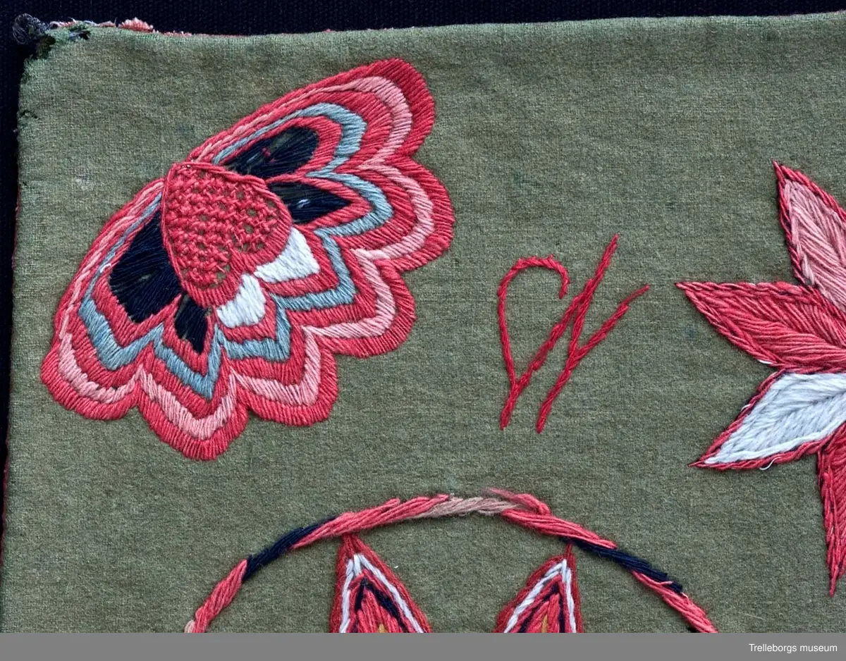 Bottentyg är grön vadmal. Tre stora ringar i kontur utgör huvudmotiv. I ringen i mitten är en stor blomma med 8 kronblad utfyllnad. I de båda ringarna vid sidan är broderat en spetsuddig stjärna med 8 vingpar. Dess centrum är en snedställd kvadrat, sydd i gallerliknande konstsöm. Utmed agedynans långsidor är broderat olika blomsterstjärnor, pionliknande blommor och en blomliknande rundning av 4 st hjärtfigurer. Färger: vitt, gult, svart, blågrått samt många olika nyanser i rött. Baksidan tegelröd yllevävnad. 
Enligt katalogen märkt I.W.D.S.  SL. Hjärtrosetter och hjärtan. 
Enligt Ernst Fischer, Årsbok från Malmö museum 1971, sid 180: En åkdyna i Tbgs museum har som mittmotiv en runduddig stjärna, omgiven av en ring samt en hjärtrosett över och under, flankerad av spetsuddiga stjärnor. T.h. och v om mittmotivet stora spetsuddiga stjärnor inom ringar. I hörnen varierande blomformer. De ganska svårtydda initialerna skulle kunna tydas som LWD IVH, ehuru bokstäverna då icke är placerade i rätt ordning. IVH skall i så fall beteckna I Vämmenhög.