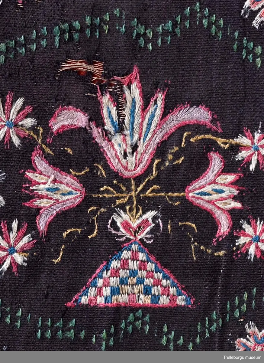 Bottenväv i svart ullgarn, rips. Varp 2 tr lingarn. Broderi och flossa i ullgarn. Runt vävnaden en bård, ca 8 cm, vävd i tränsaflossa, bestående av två blågröna kanter och mellan dessa rosa v-linje. I tränsaflossa finns dessutom: fyra hörnfigurer, timglas mitten och ytterkonturen till två ovala liggande figurer. I de ovala liggande figurerna är broderat en stor triangel, schackmönstrad, och två mindre samt dessutom en större tulpan. Svaga konturer av årtal och eventuellt initialer finns, men var alltför svårta för att tyda då dynan kom in i museets samlingar. Bakstycke av krapprött ylletyg i tuskaft, skarvat längs ena långsidan. Trasbollar i hörnen.