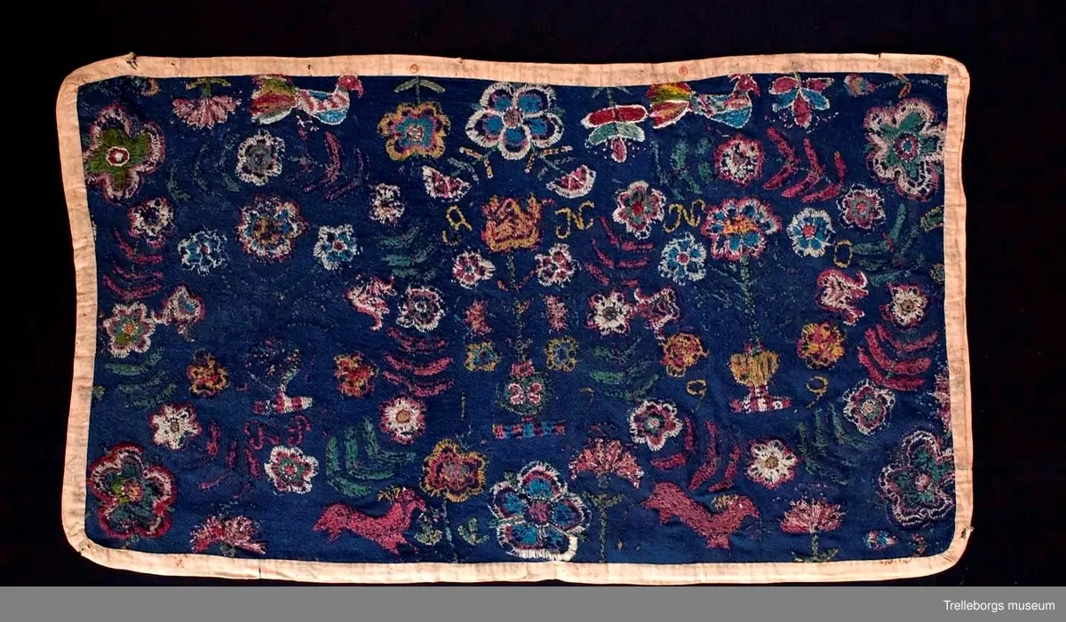 Ylletyg i tuskaft i botten, broderi 1 tr ullgarn.
Dyna av sydd flossa på blå vadmal röd kant, mönster, flera färger. Plattsöm. Anno 1799 från Gärdslöv. 
Broderiet är mkt slitet, men 2 stora kransar av blad och blommor med en blomstervas med 7 blommor i kan tydligt urskönjas. Ovanför vardera kransen finns en fågel. 
Mittfigur är en blomstervas med blommor.
Nedtill på agedynan hoppar 2 st hjortar.
Färger: mörkrött, rödlila, vitt, grönt, gult och ljusblått.
Dynan är kantad med brunt bomullstyg.
Agedynan är från Gärdslöv. 
Växter och frukter. Enligt Ernst Fischer, Årsbok från Malmö museum 1971: "Nästa åkedyna tillhör Tbgs. museum. Även den överensstämmer till alla delar med gruppens andra dynor med det undantaget, att blommorna i vasen inom kransarna är mera naturalistiskt utformade och därigenom närmar sig ursprungsdynan från 1713. Däremot är vasen mellan kransarna mer lik den föregående åkdynan.
Broderiet som är märkt ANNO 1799 och en signatur, av vilken man kan tyda IP, kommer från Gärdslöv, Vemmenhögs hd. Den bygger på samma förlaga som de övriga inom gruppen, men brodösen har icke haft samma skicklighet som de, vilka utfört dessa. Hon har förenklat teckningen och inte nyanserat detaljerna i samma utsträckning."
Sid 144-145.