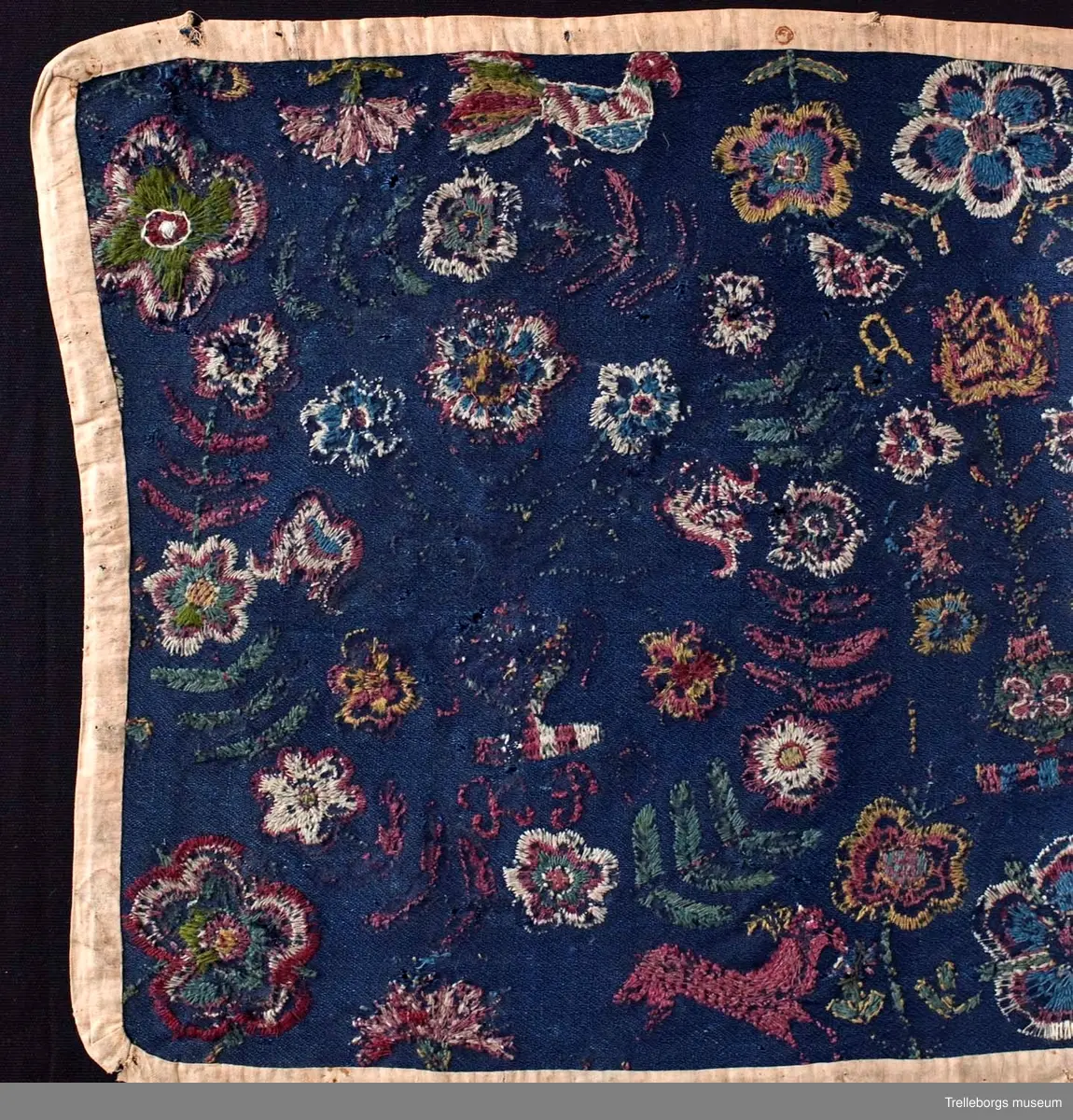 Ylletyg i tuskaft i botten, broderi 1 tr ullgarn.
Dyna av sydd flossa på blå vadmal röd kant, mönster, flera färger. Plattsöm. Anno 1799 från Gärdslöv. 
Broderiet är mkt slitet, men 2 stora kransar av blad och blommor med en blomstervas med 7 blommor i kan tydligt urskönjas. Ovanför vardera kransen finns en fågel. 
Mittfigur är en blomstervas med blommor.
Nedtill på agedynan hoppar 2 st hjortar.
Färger: mörkrött, rödlila, vitt, grönt, gult och ljusblått.
Dynan är kantad med brunt bomullstyg.
Agedynan är från Gärdslöv. 
Växter och frukter. Enligt Ernst Fischer, Årsbok från Malmö museum 1971: "Nästa åkedyna tillhör Tbgs. museum. Även den överensstämmer till alla delar med gruppens andra dynor med det undantaget, att blommorna i vasen inom kransarna är mera naturalistiskt utformade och därigenom närmar sig ursprungsdynan från 1713. Däremot är vasen mellan kransarna mer lik den föregående åkdynan.
Broderiet som är märkt ANNO 1799 och en signatur, av vilken man kan tyda IP, kommer från Gärdslöv, Vemmenhögs hd. Den bygger på samma förlaga som de övriga inom gruppen, men brodösen har icke haft samma skicklighet som de, vilka utfört dessa. Hon har förenklat teckningen och inte nyanserat detaljerna i samma utsträckning."
Sid 144-145.