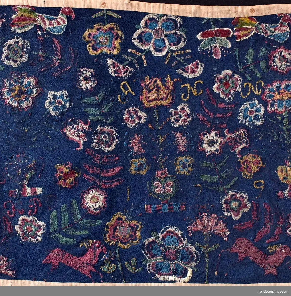 Ylletyg i tuskaft i botten, broderi 1 tr ullgarn.
Dyna av sydd flossa på blå vadmal röd kant, mönster, flera färger. Plattsöm. Anno 1799 från Gärdslöv. 
Broderiet är mkt slitet, men 2 stora kransar av blad och blommor med en blomstervas med 7 blommor i kan tydligt urskönjas. Ovanför vardera kransen finns en fågel. 
Mittfigur är en blomstervas med blommor.
Nedtill på agedynan hoppar 2 st hjortar.
Färger: mörkrött, rödlila, vitt, grönt, gult och ljusblått.
Dynan är kantad med brunt bomullstyg.
Agedynan är från Gärdslöv. 
Växter och frukter. Enligt Ernst Fischer, Årsbok från Malmö museum 1971: "Nästa åkedyna tillhör Tbgs. museum. Även den överensstämmer till alla delar med gruppens andra dynor med det undantaget, att blommorna i vasen inom kransarna är mera naturalistiskt utformade och därigenom närmar sig ursprungsdynan från 1713. Däremot är vasen mellan kransarna mer lik den föregående åkdynan.
Broderiet som är märkt ANNO 1799 och en signatur, av vilken man kan tyda IP, kommer från Gärdslöv, Vemmenhögs hd. Den bygger på samma förlaga som de övriga inom gruppen, men brodösen har icke haft samma skicklighet som de, vilka utfört dessa. Hon har förenklat teckningen och inte nyanserat detaljerna i samma utsträckning."
Sid 144-145.