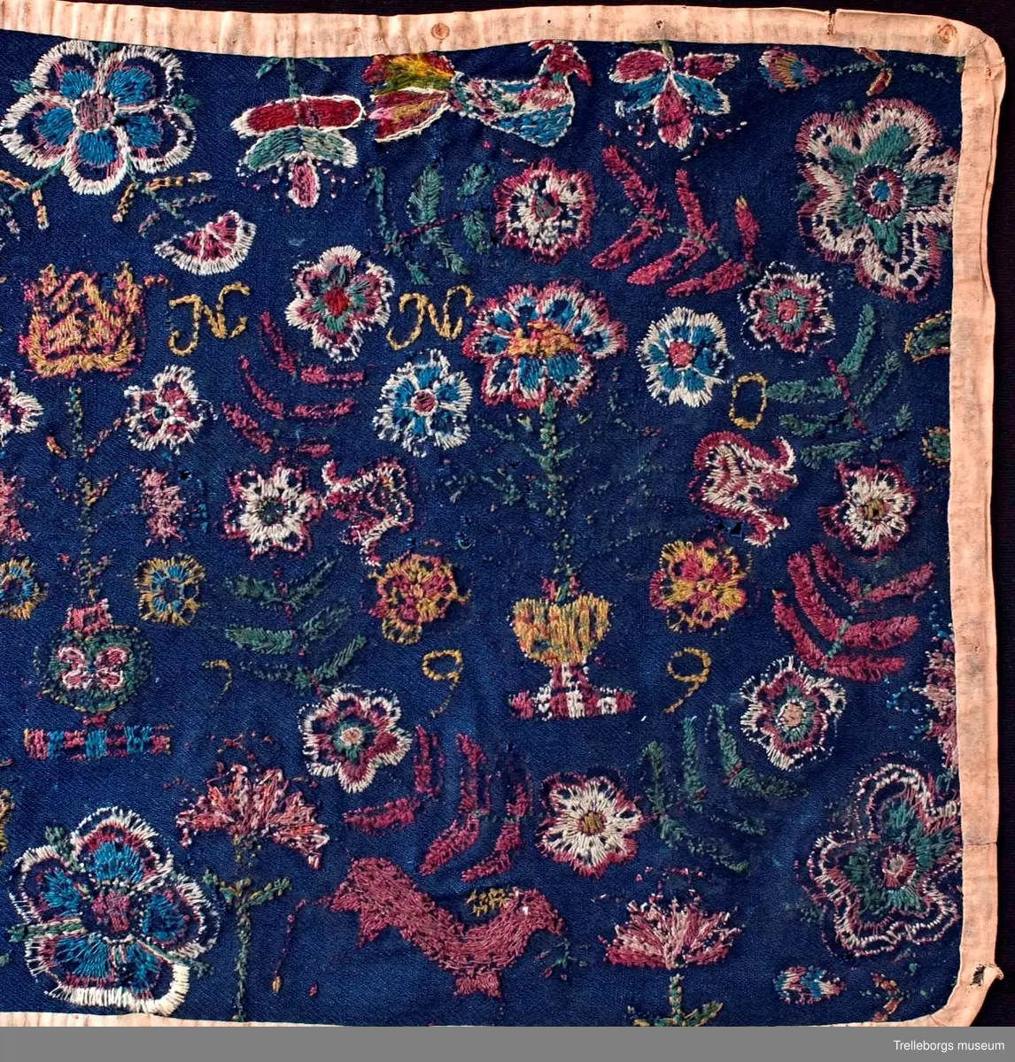 Ylletyg i tuskaft i botten, broderi 1 tr ullgarn.
Dyna av sydd flossa på blå vadmal röd kant, mönster, flera färger. Plattsöm. Anno 1799 från Gärdslöv. 
Broderiet är mkt slitet, men 2 stora kransar av blad och blommor med en blomstervas med 7 blommor i kan tydligt urskönjas. Ovanför vardera kransen finns en fågel. 
Mittfigur är en blomstervas med blommor.
Nedtill på agedynan hoppar 2 st hjortar.
Färger: mörkrött, rödlila, vitt, grönt, gult och ljusblått.
Dynan är kantad med brunt bomullstyg.
Agedynan är från Gärdslöv. 
Växter och frukter. Enligt Ernst Fischer, Årsbok från Malmö museum 1971: "Nästa åkedyna tillhör Tbgs. museum. Även den överensstämmer till alla delar med gruppens andra dynor med det undantaget, att blommorna i vasen inom kransarna är mera naturalistiskt utformade och därigenom närmar sig ursprungsdynan från 1713. Däremot är vasen mellan kransarna mer lik den föregående åkdynan.
Broderiet som är märkt ANNO 1799 och en signatur, av vilken man kan tyda IP, kommer från Gärdslöv, Vemmenhögs hd. Den bygger på samma förlaga som de övriga inom gruppen, men brodösen har icke haft samma skicklighet som de, vilka utfört dessa. Hon har förenklat teckningen och inte nyanserat detaljerna i samma utsträckning."
Sid 144-145.