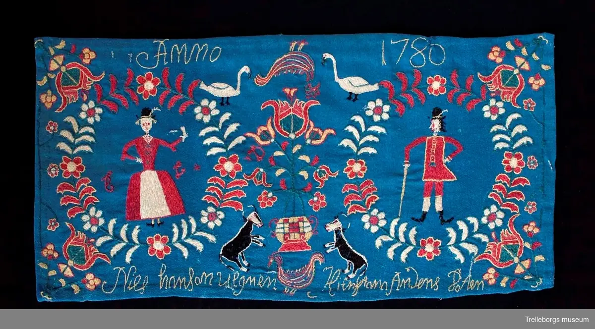 På en mellanblå botten är två kransar broderade, i den ena kransen står en kvinna och i den andra en man. I dynans hörn är blommor broderade, samma blomma kommer igen i dynans mitt tillsammans med en tupp, två gäss och två kor. Broderiets färger är rött, vitt, gult, grönt och svart. Dynan är märkt Anno 1780 Nils Hansson Ulgren Kierstin Andersdotter.