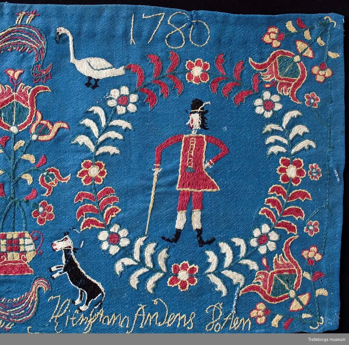 På en mellanblå botten är två kransar broderade, i den ena kransen står en kvinna och i den andra en man. I dynans hörn är blommor broderade, samma blomma kommer igen i dynans mitt tillsammans med en tupp, två gäss och två kor. Broderiets färger är rött, vitt, gult, grönt och svart. Dynan är märkt Anno 1780 Nils Hansson Ulgren Kierstin Andersdotter.