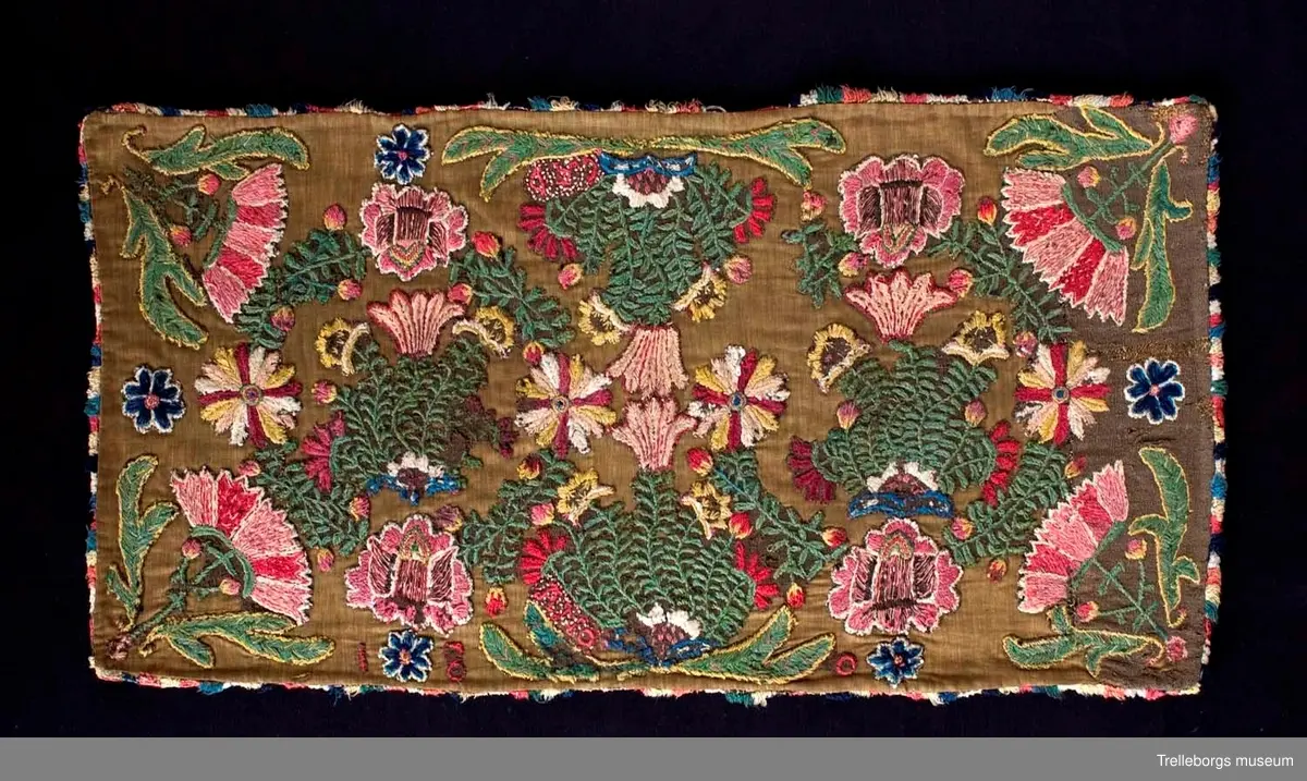 Broderad agedyna på grov, brun yllebotten. Motivet är två grönskande blomsterkransar fyllda med vad som mest liknar blommande buskar, även mellan kransarna finns två blommande buskar. Här är dynan märkt med CAD i den ena och ANO i den andra, båda på ett litet fält med franska knutar i vitt och rött. Under busken med ANO står 180 ganska glest, sista siffran finns inte kvar. Dynan är monterad med en flerfärgad kavelfrans och en klarröd, ganska grov yllebaksida.