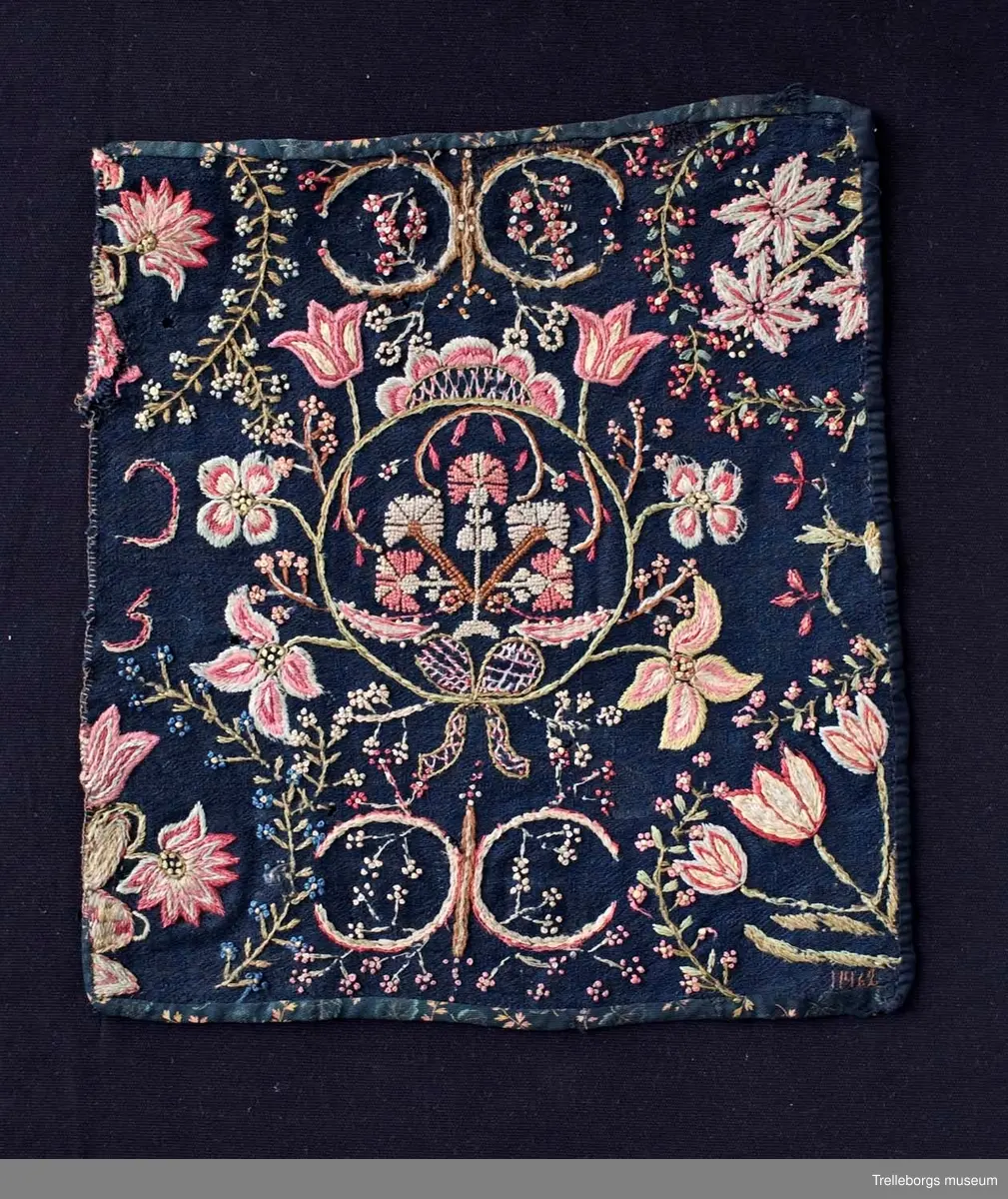 Botten svart ylletyg i tuskaft, broderi: 2 tr ullgarn. Del av åkdyna i plattsömsbroderi, schattersöm. Dynans sirliga rikt utformade mönster är mycket ovanligt. Färgerna är ljusa i rött, rosa, blått och gult. På mitten är en rund ring broderad, denna har en mittfigur sydd med små korsstygn. Runt ringen finns blommor typiska för schattersöm. I varje hörn finns tulpanliknande blommor omgivna av en halv krans småblommor. Småblommorna förekommer över hela ytan. Blomsterbuketten i mitten är sydd i små korsstygn, rosetten i bottensöm, de mindre blommorna är sydda med knutar. Kantad med tyg. Endast halva dynan. Se även Årsbok för Malmö museum sid 44-45, Ernst Fischer: I Trelleborgs museum finnes hälften av en ursprunglig åkdyna som har ett mittmönster av en ring-krans med ändarna nedtill sammanknutna av ett band med ögla samt krönt av ett ornament. Från kransen utgår upptill två tulpaner, på mitten två fyrbladiga blommor, och nederst två utslagna tulpaner. Mellan dessa blommor små kvistar. Inom kransen en växt med fem nejlikor sydda i korssöm. I övre hörnet en växt med fem nejlikor sydda i korssöm. I övre hörnet en växt med sexbladiga blommor och i det nedre en tulpanstjälk med tre blommor. Båda omramade av en spröd kvist. Till vänster om kransen upptill och nedtill hälften av en urna med tre stora blommor, vända mot varandra. Även dessa omramade av liknande kvistar.
