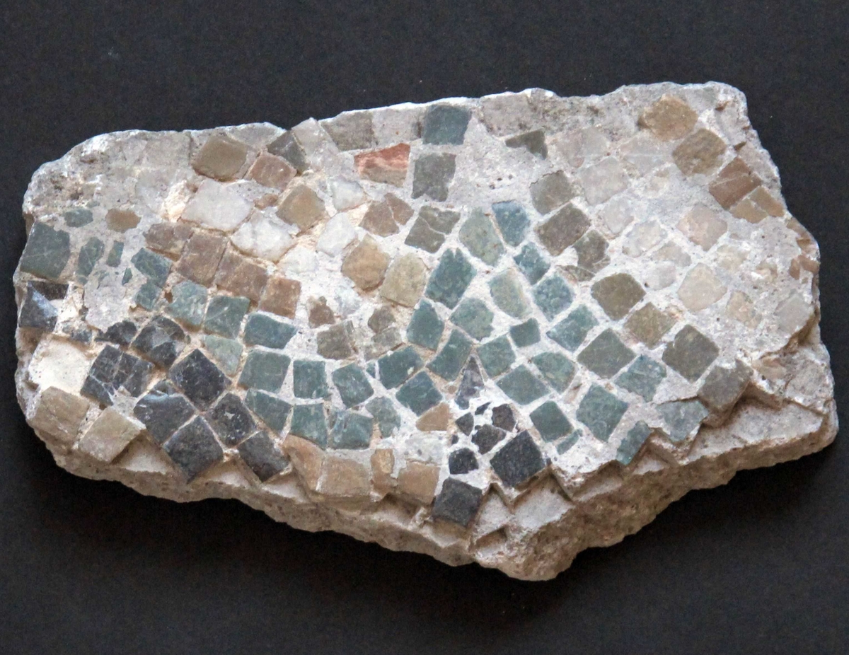 Mangefarget mosaikk fragment, tilnærmet rektangulært av form. Grønne, hvite, grå og gule tesseraer i naturstein, satt i fin kornet, hvit mørtel. Steinene av ulik størrelse, satt bueformede mønster.