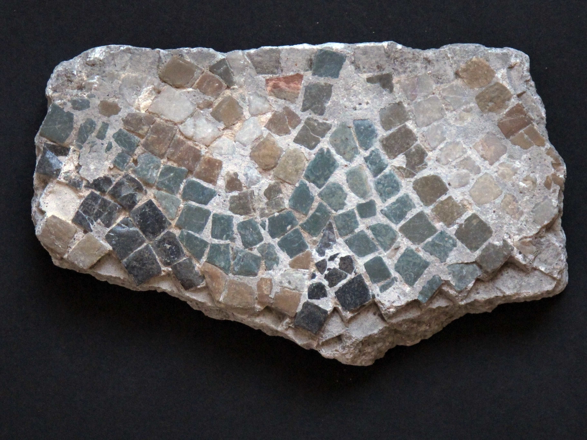 Mangefarget mosaikk fragment, tilnærmet rektangulært av form. Grønne, hvite, grå og gule tesseraer i naturstein, satt i fin kornet, hvit mørtel. Steinene av ulik størrelse, satt bueformede mønster.