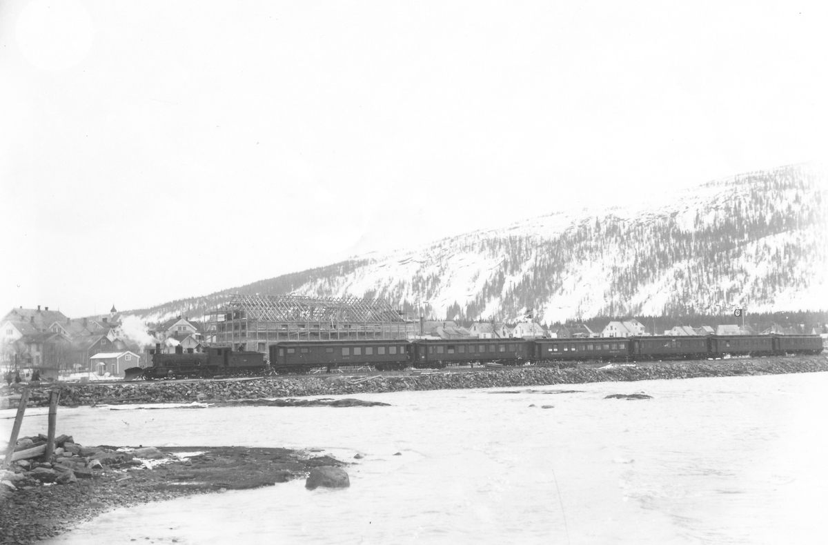 Tog med vogner kjører nordover. Mo jernbanestasjon under oppførelse. Foto tatt fra sjøsiden.