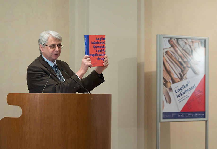 Prof. Dr. Jacek Purchla, direktør ved ICC, står bak talerstolen og holder opp utstillingskatalogen på åpningen av Logika Lokalnosci, 16. desember 2016. Plakaten for utstillingen sees i bakgrunnen.
