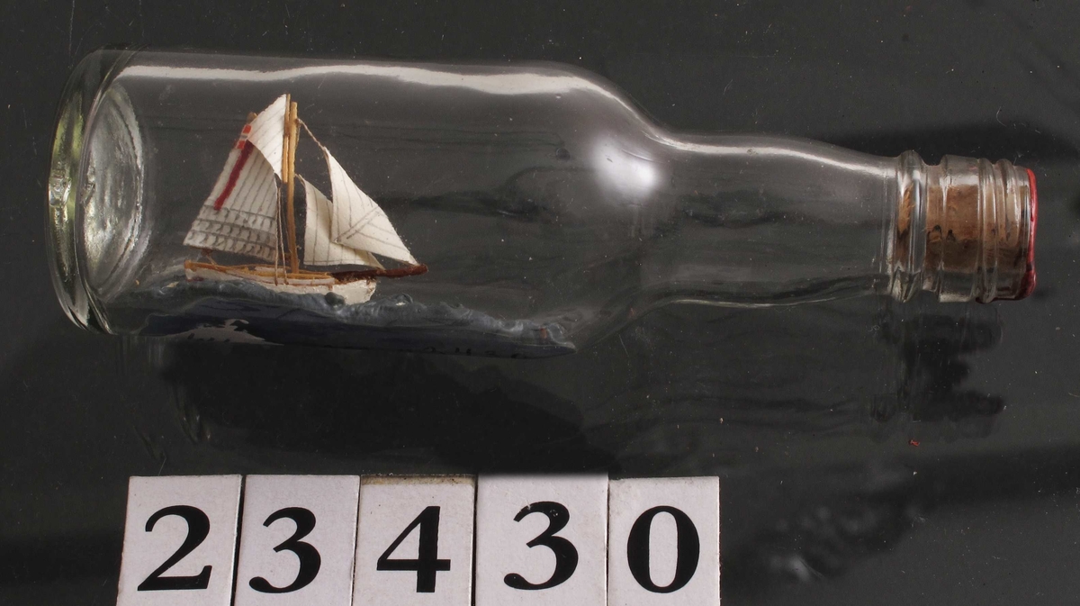 Mini flaskeskip: Losbåt.  Glass, klart. Kork med rød lakk. Hvitmalt miniatyrskute med rød bunn og rød stripe i seilet. Blå sjø.