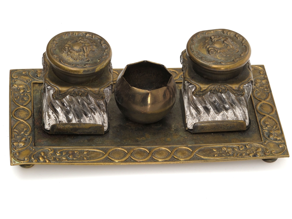 Skrivestell. Presset  messing, 2 blekkhus av  pressglass. Rektangulær messingplate med presset ornamentbord langs kanten og 4 små dreide føtter. Presset dekorativt mønster.
