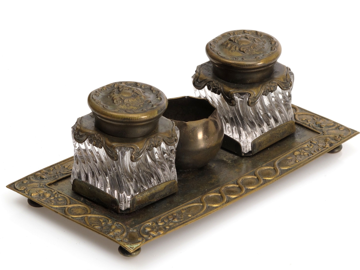 Skrivestell. Presset  messing, 2 blekkhus av  pressglass. Rektangulær messingplate med presset ornamentbord langs kanten og 4 små dreide føtter. Presset dekorativt mønster.